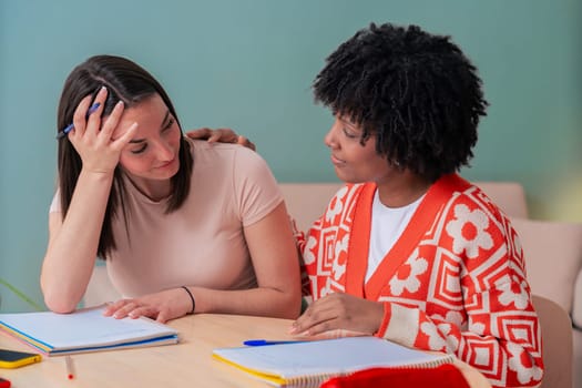 Two multiracial female friends studying at home. One appears stressed while the other offers support with a hand on her shoulder. Emotional moment of empathy, care, and friendship during learning.