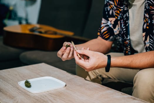 Asian young man sitting on a couch with a cannabis bud in his hand and smoking a joint relaxing time at home. A guitar is in the background