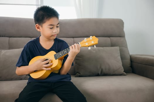 Cheerful boy in a blue shirt plays a ukulele on a modern couch in a sunny living room. The scene captures the spirit of creativity and relaxation in a family-friendly environment.