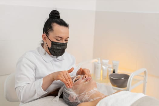 Middle-aged woman undergoing facial care in cosmetology clinic. Beautician applies gauze on moisturized skin with serum to begin carboxytherapy, promoting hydration, glow, tone, rejuvenation effect.