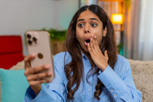 Indian woman receives surprising good news on smartphone while sitting on home sofa, reacts with wow expression. Arabian girl opens mouth in shock, shows hand gesture with amazement and emotion.