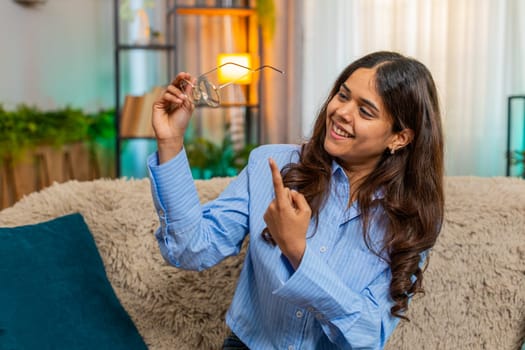 Indian woman laughs while searching for glasses on home sofa. Arabian girl shows silly joy, confusion and fun in humorous personal moment with light-hearted emotion.