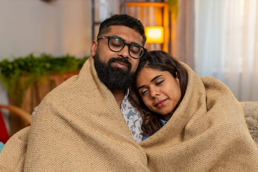 Indian couple man and woman sitting on home sofa, sharing soft hug under warm blanket in silence and comfort. Young family enjoying peaceful cozy moment together, feeling safe and emotionally close.