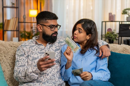 Indian couple man and woman sitting on home sofa holding money dollar cash and smartphone while planning purchases and family budget. Discussing future expenses, savings and income from salary indoors