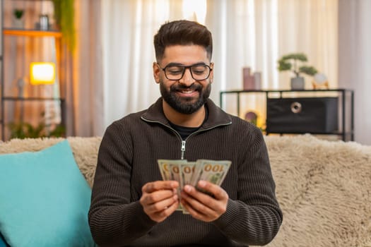 Indian man at home happily counts cash dollars while sitting on sofa, enjoying recent income or lottery win. Arabian guy expresses joy and financial confidence, celebrating success with satisfaction.