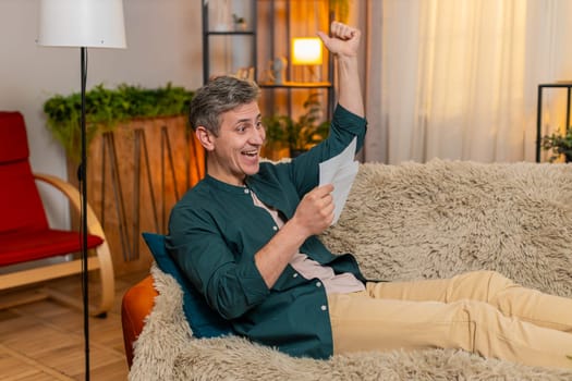 Young happy man at home opens letter and happily celebrates great news, raising fist with joy. Caucasian guy on sofa feels excited about job offer, loan approval or successful school exam result.