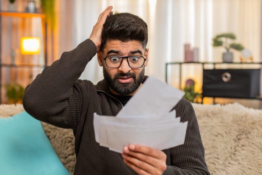 Indian man at home checks bills receipts with tense expression, nervously grabbing his head in frustration. Arabian guy on sofa struggles with financial pressure and visible signs of emotional stress