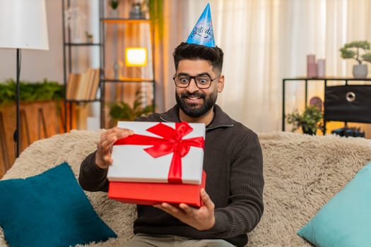 Indian man at home happily opens birthday gift while sitting on sofa with smile and excitement. Arabian guy looks surprised and joyful, thrilled by unexpected present and special celebration moment