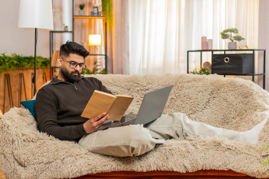 Indian man at home studies with serious look, flipping textbook pages beside laptop with concentration. Arabian guy on sofa prepares for exam with tension, focused on remembering all key material.