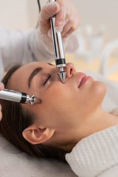 Caucasian girl lies relaxed during forehead zone rejuvenation with microcurrent device. Young woman gets facial procedure from cosmetologist in beauty clinic, enhancing skin tone and freshness.