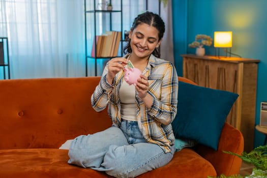 Indian young woman thoughtfully counting cash dollar bills, placing money into piggy bank at home. Arabian happy girl focused on saving, budgeting carefully, experiencing limited financial resources