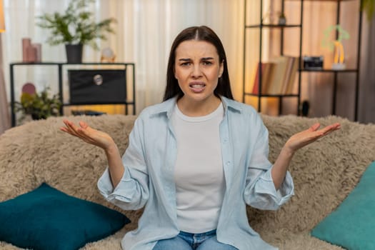 Young confused woman at home on sofa making surprised What Why facial expression looking shocked, confused, puzzled. Caucasian girl emotionally reacting, expressing astonishment and sudden disbelief.