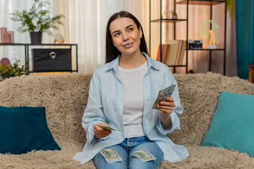 Young woman at home counting cash dollars thoughtfully reviewing her budget, sighing and setting money aside. Caucasian girl on sofa carefully managing personal finances, expenses and savings, salary