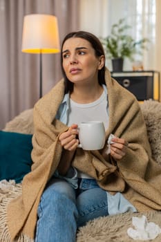 Young woman at home wrapped warmly in plaid blanket holding hot tea sitting on sofa feeling sick. Caucasian girl experiencing cold symptoms, resting, comforted by warmth of beverage, unwell mood