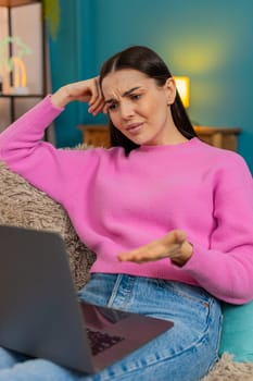 Young woman at home dissatisfied looking at laptop screen leaning head on hand tiredly with upset expression. Caucasian girl on sofa feeling fatigue, frustrated from workload and negative emotions