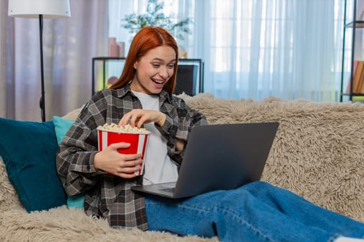 Happy woman at home watching movie on laptop and eating popcorn while laughing with joy and excitement. Caucasian redhead girl on sofa enjoying emotional scene, relaxed evening with fun entertainment