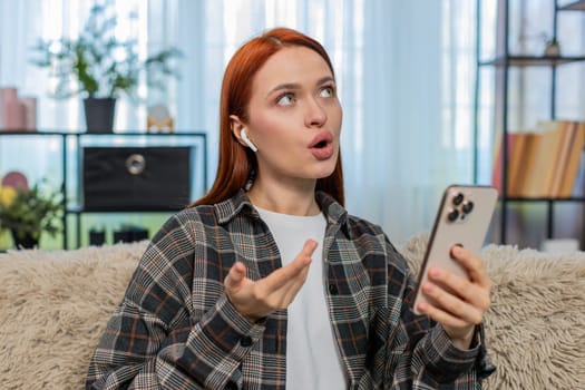 Young woman at home learning new language using mobile app and repeating words aloud with focus and joy. Caucasian redhead girl on sofa enjoying educational process practicing pronunciation and speech