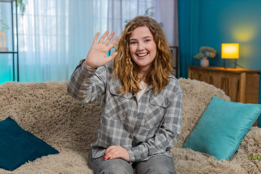 Young woman at home smiling warmly and waving hand to say hello with friendly and cheerful energy. Caucasian redhead girl on sofa expressing joy, welcoming gesture and positive emotional connection