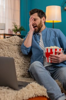 Adult man at home enjoys popcorn while watching movie on laptop, fully immersed in scene. Caucasian guy laughing in excitement at an unexpected moment, reacting emotionally to the thrilling storyline