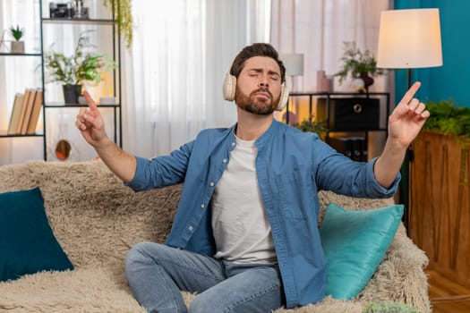 Young man at home wearing headphones raises hands as if conducting an orchestra, fully enjoying the music. Caucasian guy on sofa moves rhythmically, lost in melody, expressing passion and creativity
