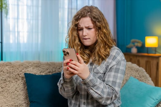 Young woman at home angrily texting on mobile phone reacting with irritation and frustration to message. Caucasian redhead girl on sofa waves hand annoyed, emotional expression of dissatisfaction