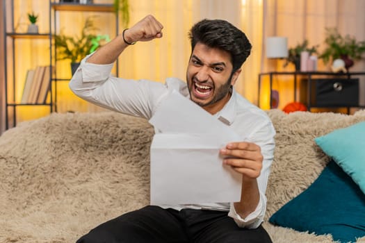 Indian man at home reading a letter with great news, celebrating a job offer, positive test results, or lottery win. Arabian guy expressing joy and excitement, celebrating success with a big smile