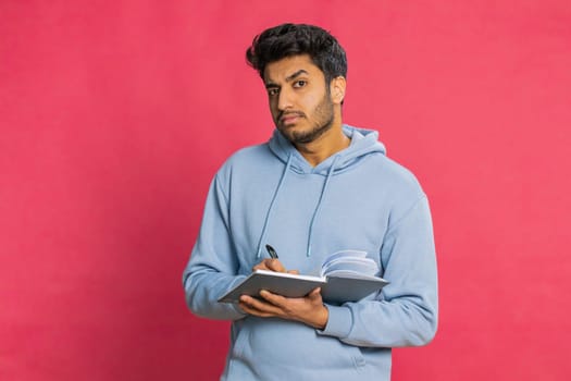 Thoughtful journalist young Indian man making notes, writing down thoughts with pen into notepad notebook diary, to do list, good idea inspiration. Arabian Hindu guy isolated on red background