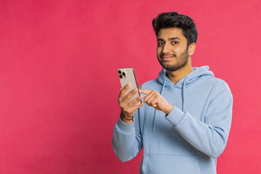 Happy Indian young man browsing social media, smiling while looking at smartphone, enjoying interesting content and typing a message. Relaxed Arabian guy engaging online, isolated on red background
