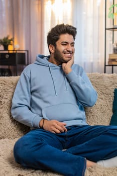 Dental problems. Young Indian man touching cheek, closing eyes with expression of terrible suffer from painful toothache, sensitive teeth, cavities. Hindu guy at home apartment living room on sofa.