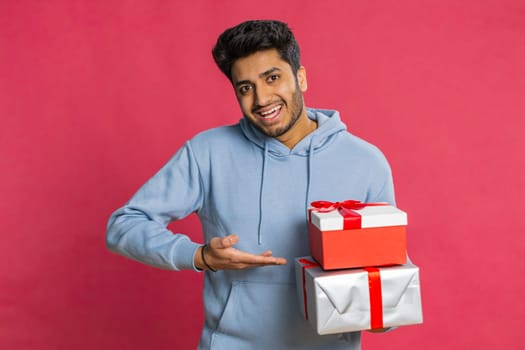 Positive smiling Indian man presenting birthday gift boxes stretches out hands, offer wrapped present career bonus, celebrating party promotion discount sale. Arabian guy isolated on red background