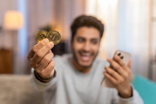 Young Indian man with bitcoin checks cryptocurrency exchange charts on smartphone app calculates approximate profits, looking on BTC coins in hand celebrating win. Hispanic guy looks satisfied at home