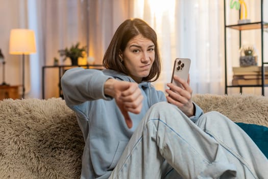 Dislike. Upset Caucasian woman using smartphone sitting on sofa couch at home showing thumbs down sign gesture, expressing discontent, disapproval dissatisfied bad work. Displeased girl shakes head.