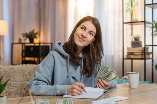 Focused young Caucasian woman making shopping list in notebook sitting at table on sofa looking at receipts and counting money in living room at home. Girl preparing notes for financial cost or budget
