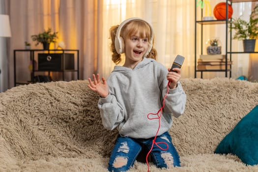 Young teenager girl wears headphones at home dancing, humming a favorite song while holding toy microphone. Blonde child kid on sofa waves hands, picturing grand performance and personal mini concert