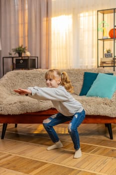 Healthy teenager girl stands at home performing simple squats and counting one, two, three with growing effort. Child kid sits on sofa finally collapses in laughter feeling happy after a brief workout