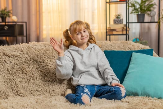 Young teenager girl waving hand with happy smile at home, expressing joy and friendliness. Caucasian child kid sitting on a sofa, saying hello hi welcome or goodbye with cheerful and relaxed attitude