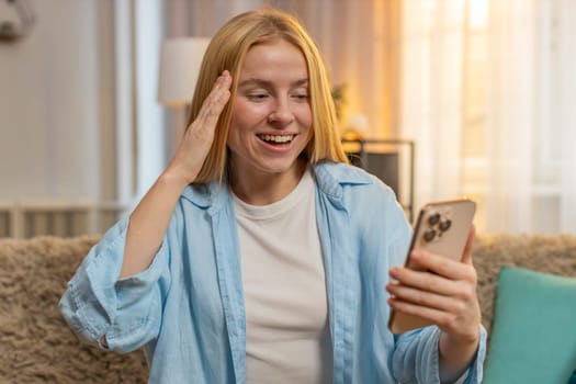 Young woman sits at home checking smartphone, shouting excitement after receiving good news and feeling triumphant. Caucasian girl jumps on sofa, showing victory gesture celebrate win, proud and happy