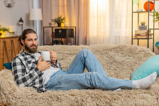 Young man resting lying at home, holding warm cup of coffee or tea with gentle grin and eased shoulders. Caucasian guy on sofa feels soothing quiet, appreciating short pause from busy tasks and noise.