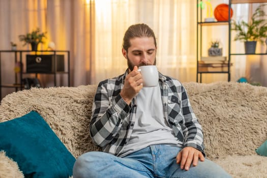 Young man holding steaming coffee or tea at home, closing eyes to soak smell in comforting fragrance. Guy on sofa inhales deeply, relishing each slow drinking sip mouthful with visible satisfaction.