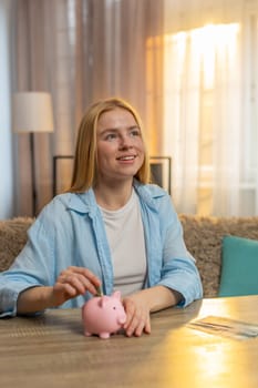 Young woman stores cash at home, dropping bills into piggy bank with contented grin and hopeful thought. Happy Caucasian girl sits at table radiates satisfaction aiming for future needs or investments