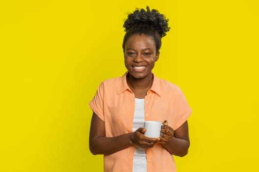 African American woman holding white cup near face, breathing in soothing scent with smile of appreciation. Black girl on yellow background showing comforting relaxation, drinking morning hot coffee