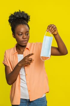 African American woman holds a medical protective mask in one hand while gesturing, explaining how its use. Black girl on yellow background shows focus and interest in discussing health and safety