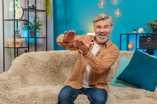 Caucasian mature man sitting on home sofa, holding imaginary money cash and making a giving away gesture. Middle-aged guy looks happy, distributing wasting money, generously donating, sharing profits