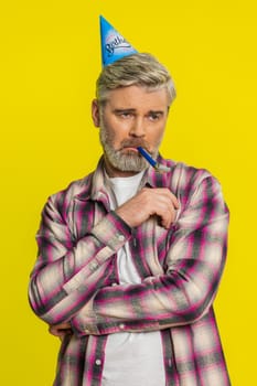 Mature man stands alone with party birthday cone hat and whistle blower showing dull spirit, disappointment. Middle-aged guy on yellow background feeling abandoned lonely, upset, tired, bored, unhappy
