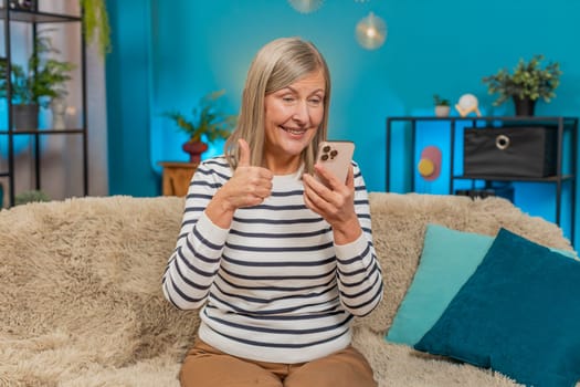 Caucasian mature woman smiles, engages with social media photos videos on smartphone while sitting at home. Grandmother gives thumbs up gesture expressing approval and happiness on sofa in living room