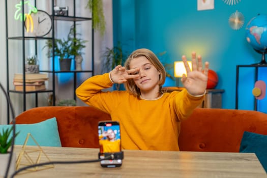 Caucasian teenager boy creating trendy selfie videos on smartphone at home. Performing dance moves while sitting on couch by table, engaging in social media content with enthusiasm and creativity
