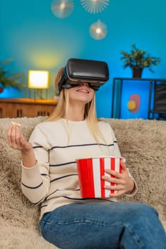 Woman wears VR headset watching movie eating popcorn at home on sofa. Girl looks excited, surprised, joyful entertainment indoors laughing cheerful emotion modern lifestyle fun digital streaming cozy