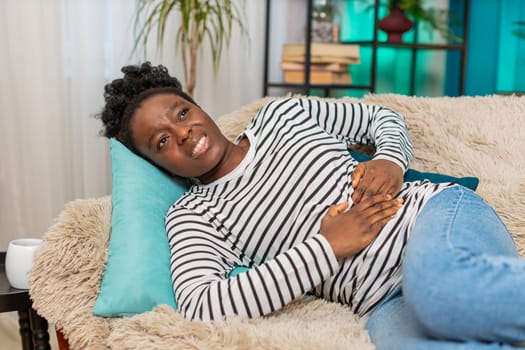 Black African American young woman suffering intense stomach pain, holding abdomen, lying on sofa at home. She looks distressed, tired, unwell, struggling with discomfort in cozy apartment living room