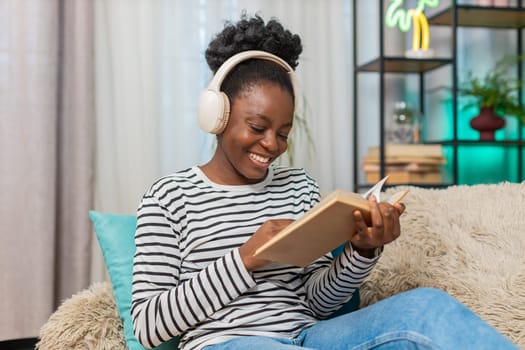 African American young adult woman reading novel at home. Sitting on sofa, wearing headphones, enjoying music, immersed in book. Relaxed atmosphere, quiet space. Living room interior, indoor leisure.