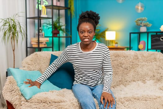 African American woman expressing anger. Sitting on home sofa, punching pillow with frustration, intense emotional reaction, showing stress or irritation releasing emotions through physical action